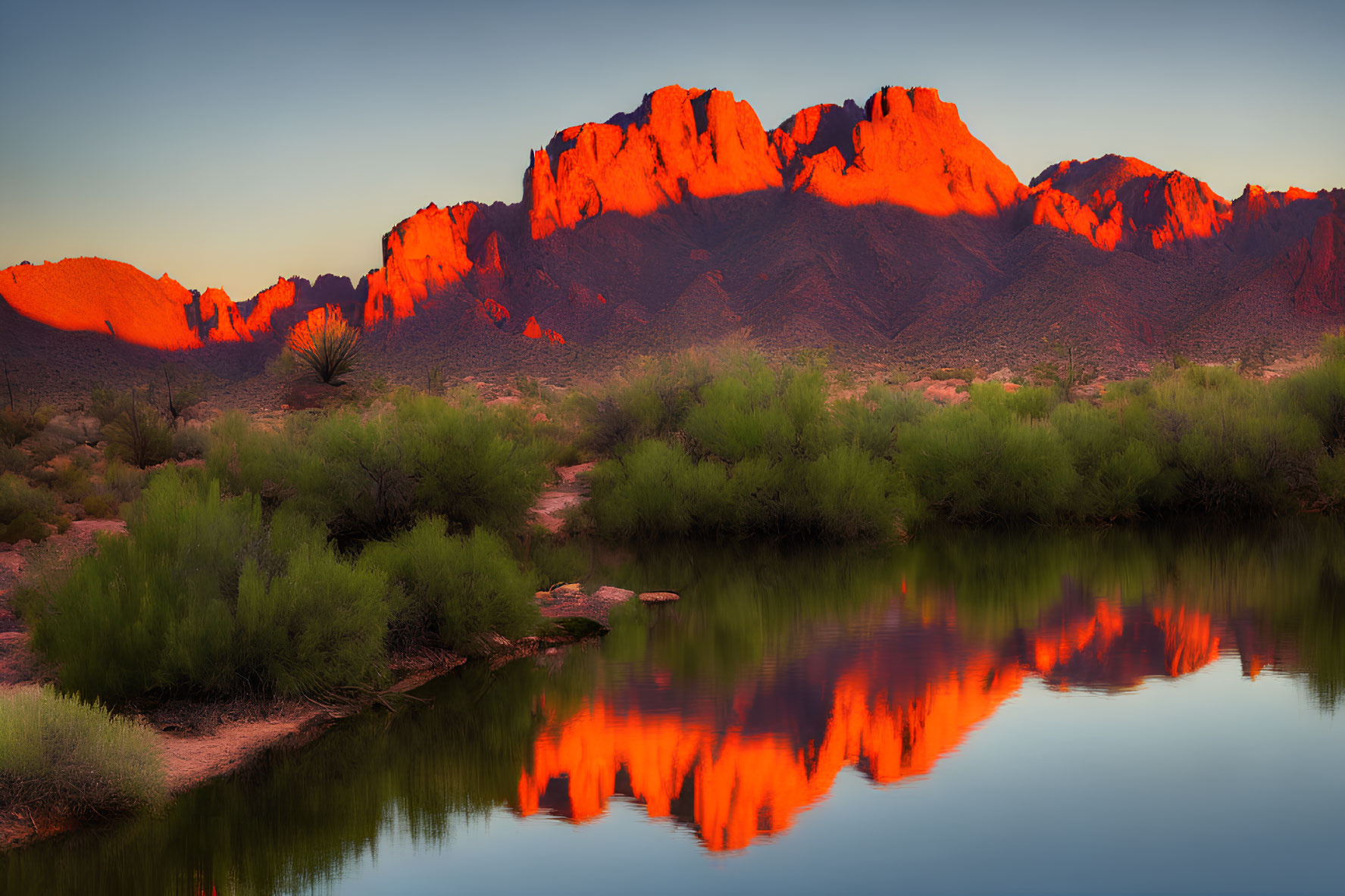 Vibrant sunset over mountain peaks, reflecting river, and desert vegetation.