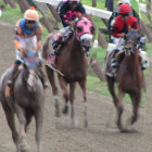 Colorful jockeys on racehorses compete in close muddy race.