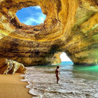Person admires sea cave with bridge on sandy beach.