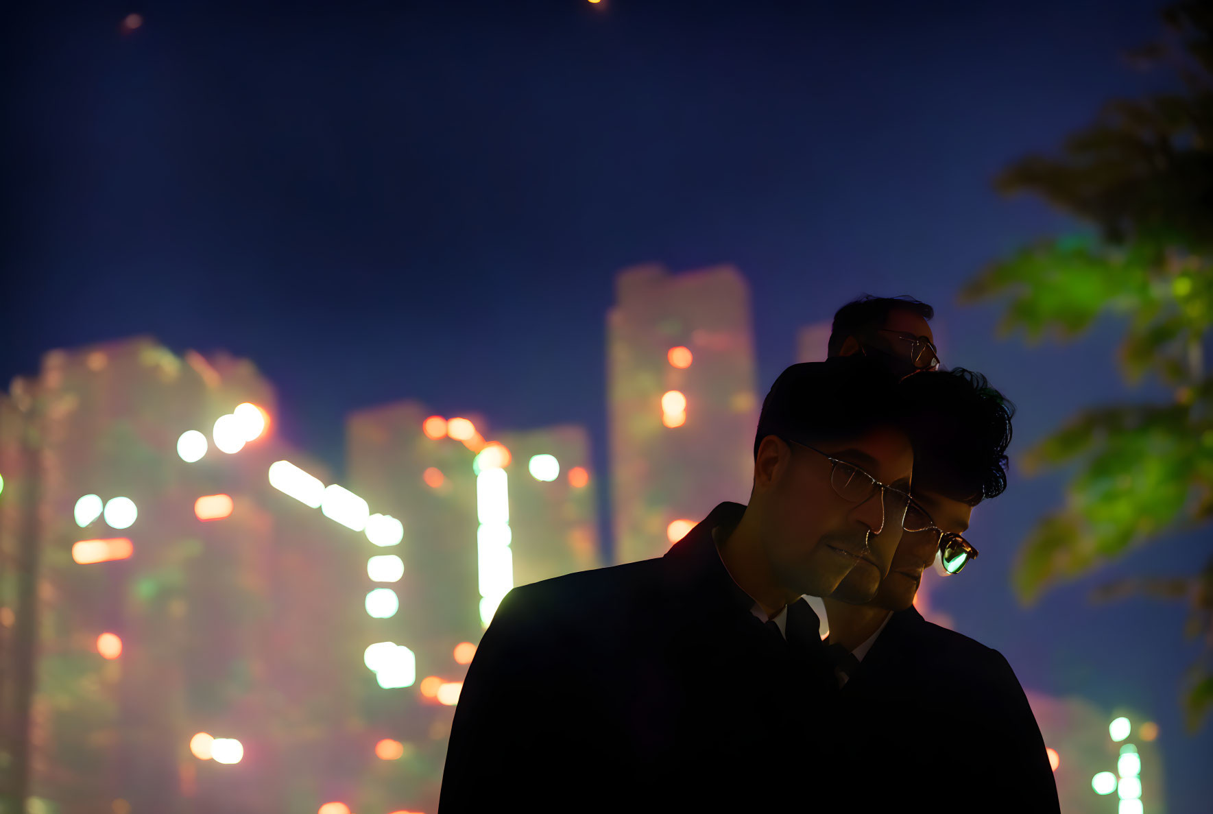 Man in glasses and dark attire stands at night with city lights bokeh.