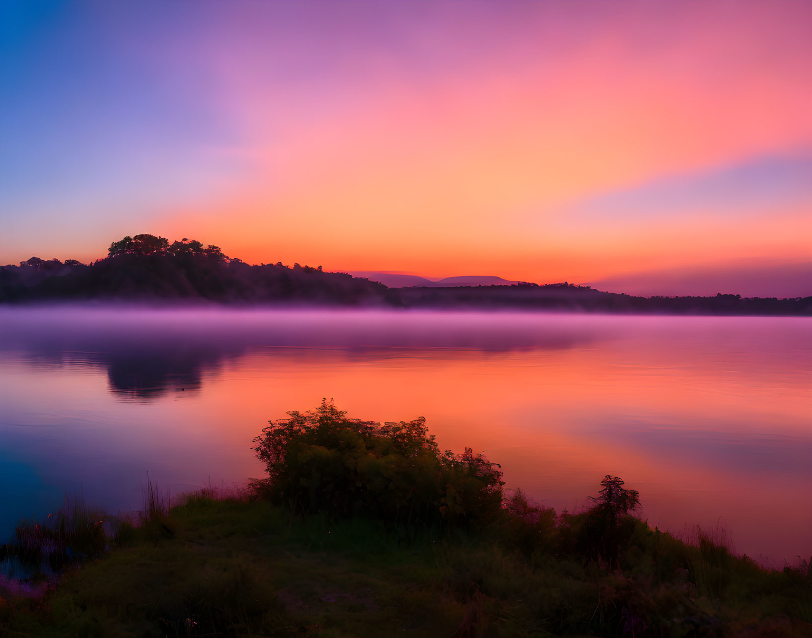 Tranquil lake at sunrise with mist, silhouetted hills, and foliage