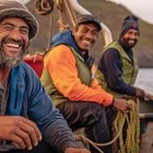 Three weathered fishermen at sea in rugged attire on a wooden boat against rocky cliffs.