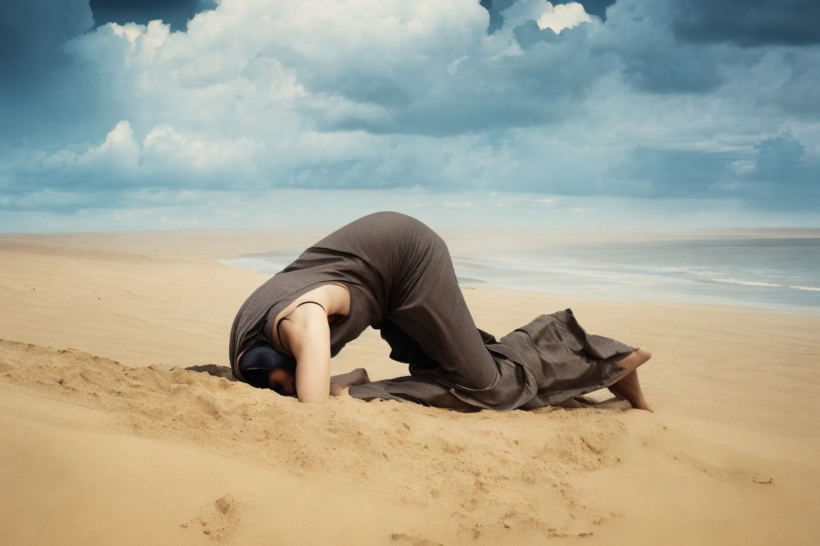 Person in grey outfit doing child's pose on sandy beach under cloudy sky