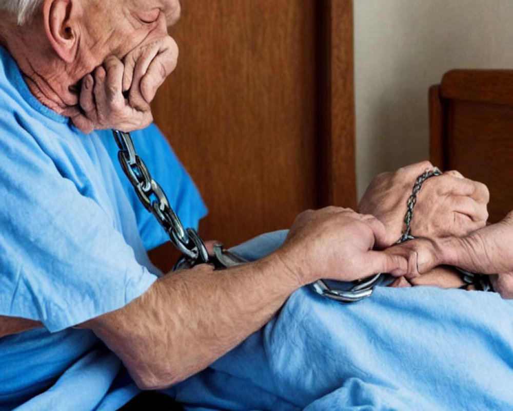 Elderly person in blue outfit handcuffed, sitting on bed distressed