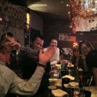 Three people at a vibrant bar with large cat figurine, man touching cat's paw to friend's