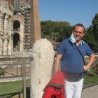 Man smiling next to classical sculpture in park with white statues and trees under blue sky