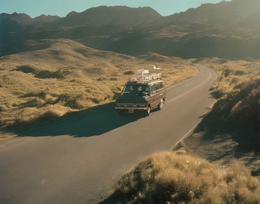 Vintage car with luggage on roof rack driving on winding road amidst grassy hills and mountains.