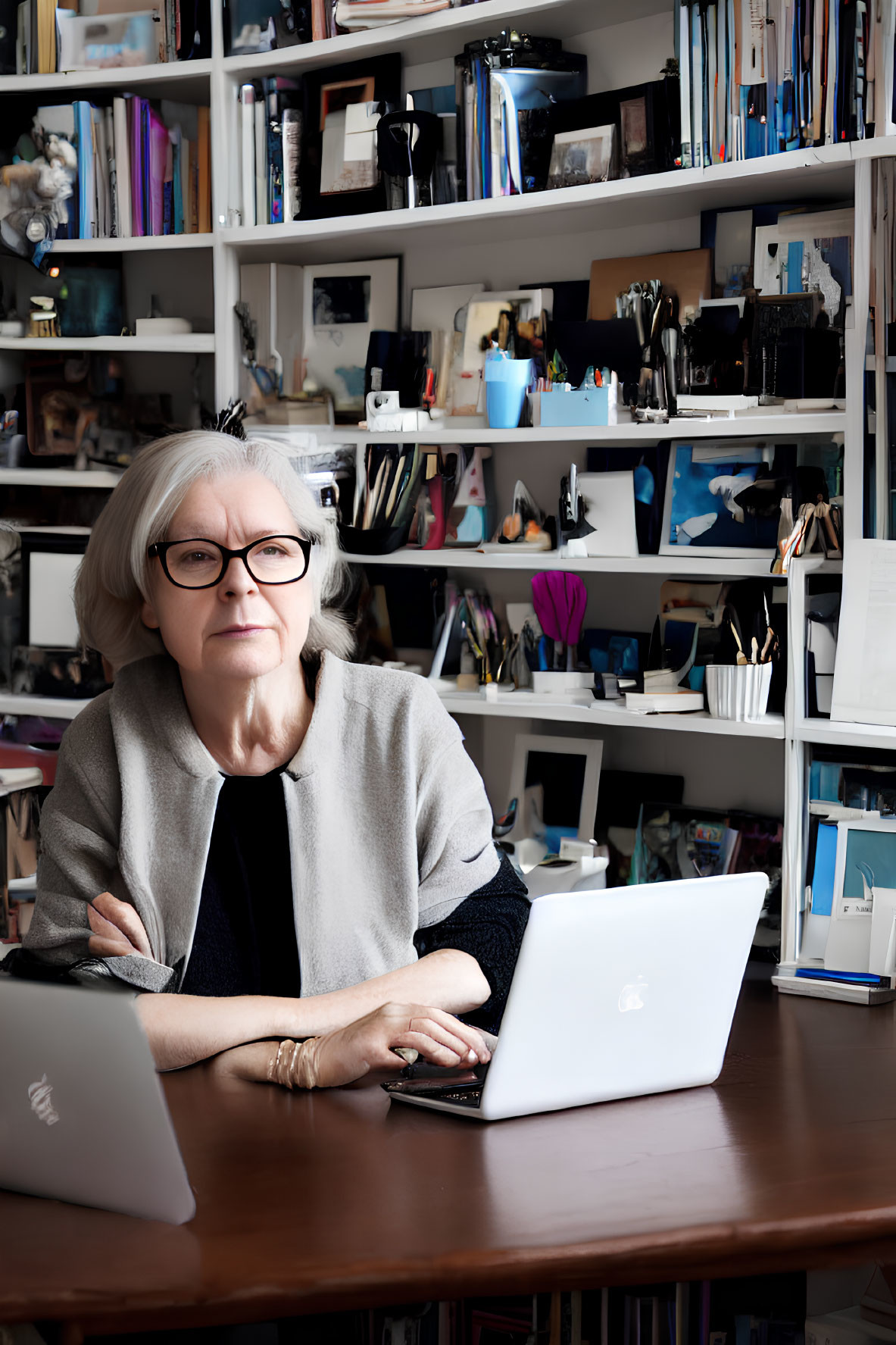 Silver-haired woman with glasses at desk surrounded by books & office supplies