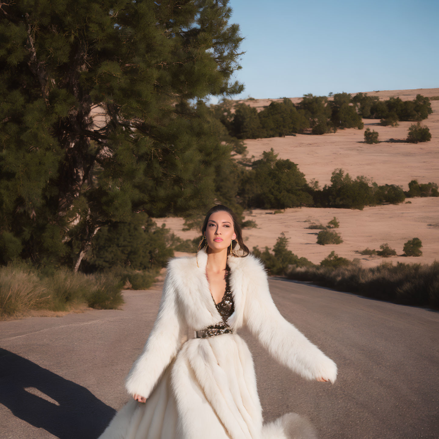 Woman in white fur coat on road with trees and sandy hills at golden hour