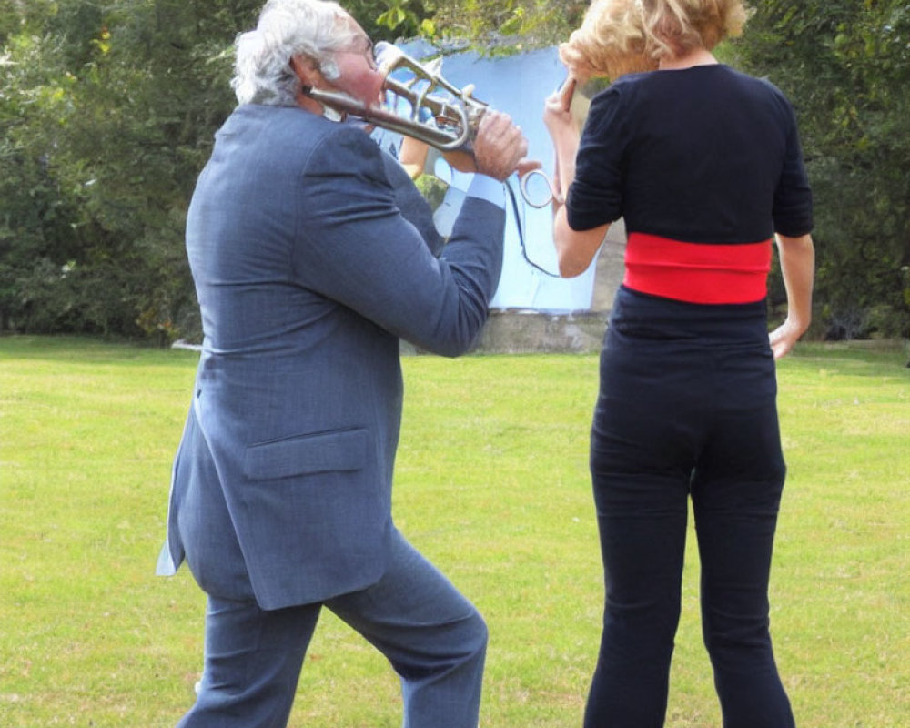 Two people on grass, one playing trombone near other person's ear.