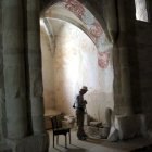 Person in historical clothing at sunlit archway entrance to tranquil forest surrounded by ancient walls.