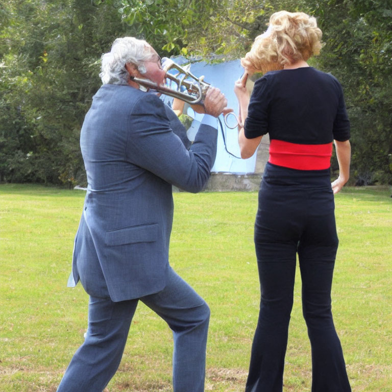 Two people on grass, one playing trombone near other person's ear.